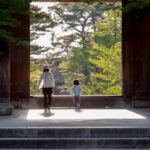 A family strolls through a temple gate surrounded by lush greenery in Kyoto, Japan.